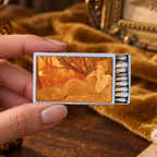 A hand holds a vintage-style matchbox with a warm-toned illustration of a reclining woman on the front, set against a styled background with velvet fabric, dried ferns, and antique decor.