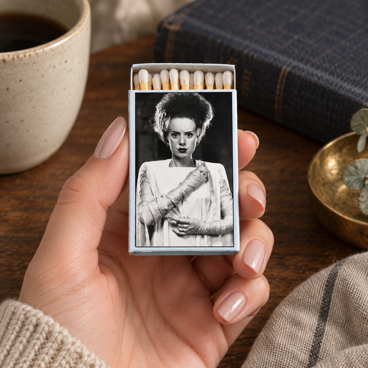 A hand holds a matchbox featuring a black-and-white portrait of a woman in vintage clothing. The matchbox is surrounded by a warm tabletop setting with a cup of coffee, a book, and a small dish.