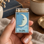 A hand with manicured nails holds a matchbox featuring a stylized moon illustration labeled 'LA LUNA' and the number '23'. The matchbox is shown in a styled tabletop setting with a book, ceramic cup, and fabric background.