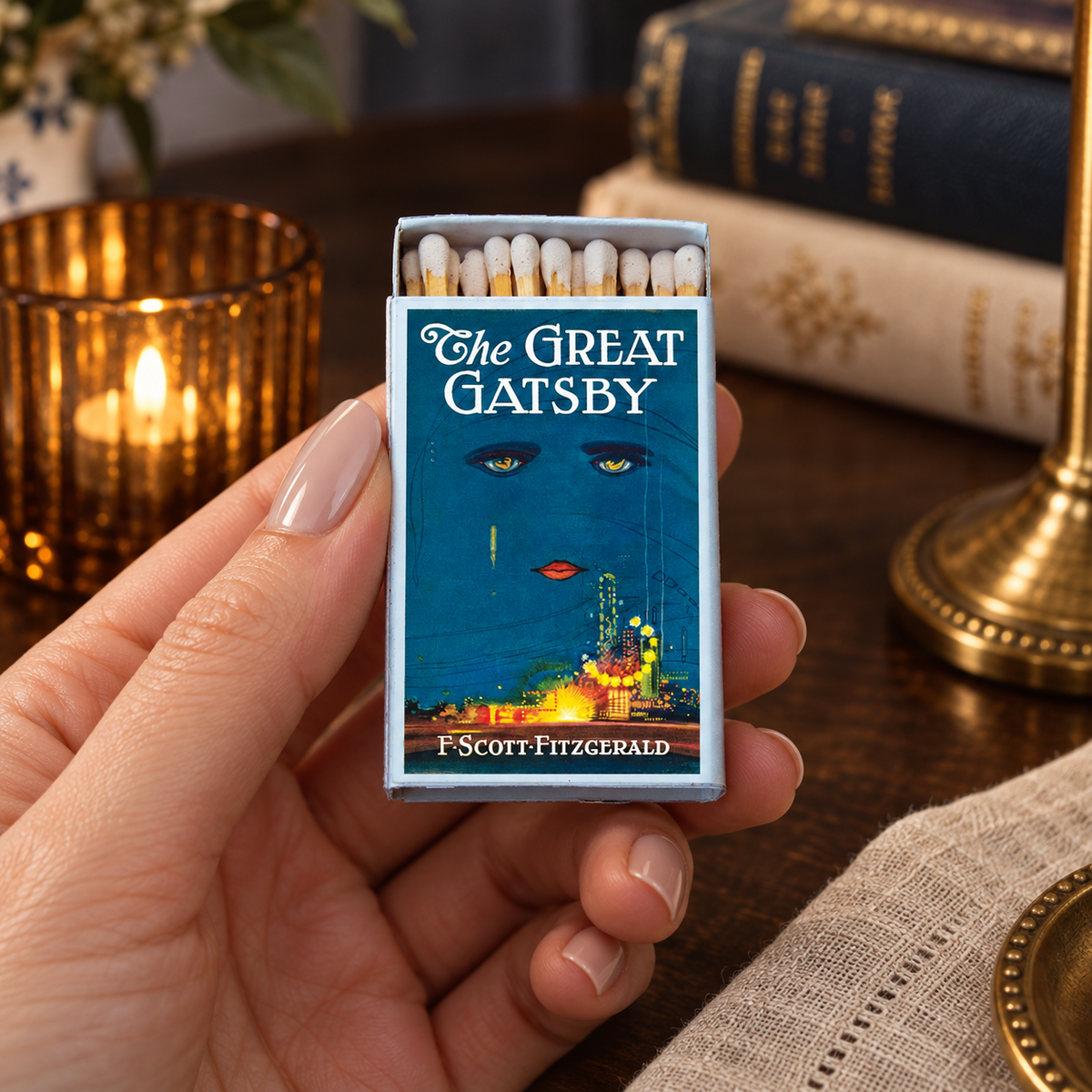A hand holds a matchbox styled like a book cover for F. Scott Fitzgerald's 'The Great Gatsby'.