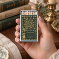 A hand holds a vintage-style matchbox with a dark green and gold peacock pattern and the text 'PRIDE PREJUDICE'. The background shows a wooden table with antique books and a brass candlestick holder.