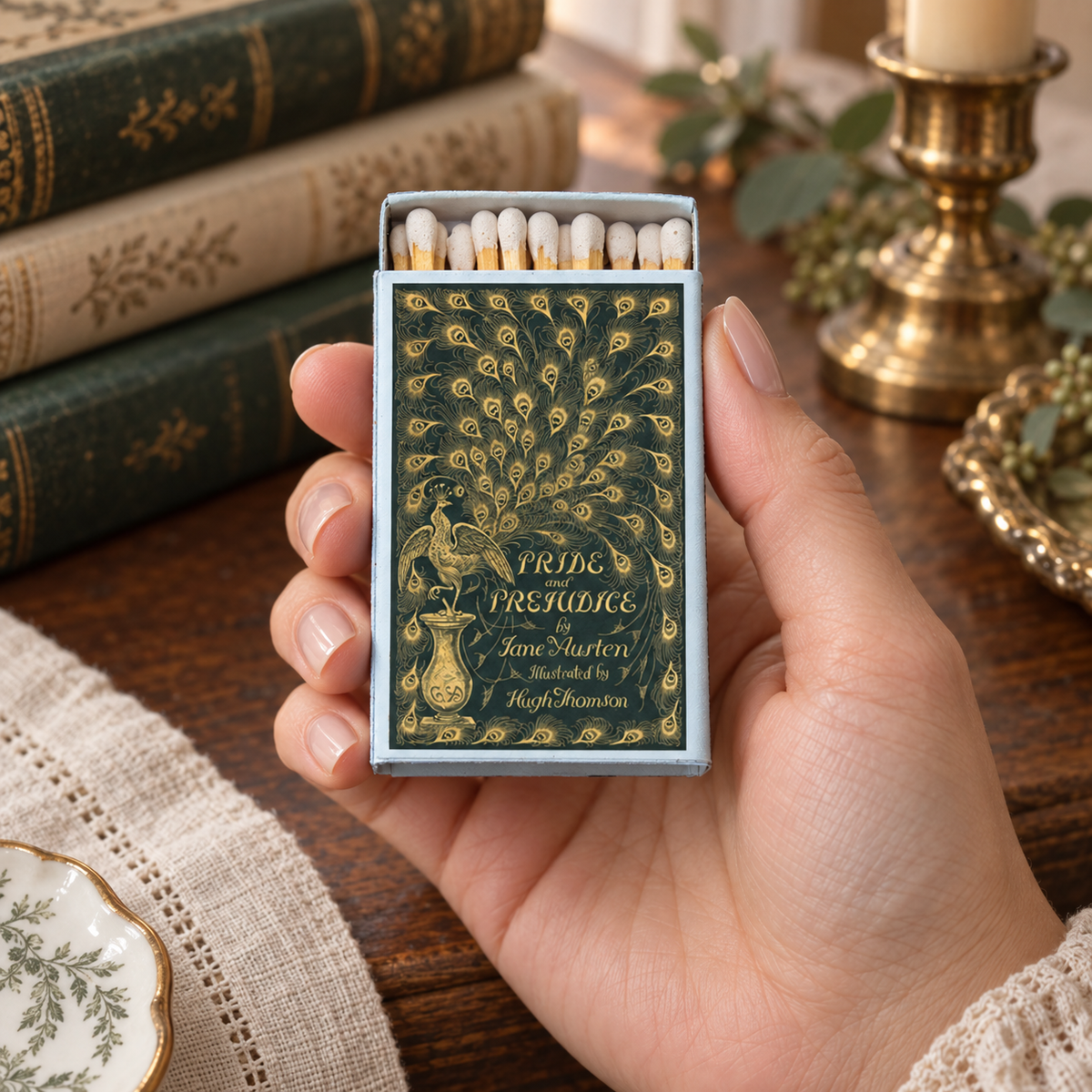 A hand holds a vintage-style matchbox with a dark green and gold peacock pattern and the text 'PRIDE PREJUDICE'. The background shows a wooden table with antique books and a brass candlestick holder.