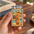 A hand holds a matchbox decorated with a Van Gogh-style sunflower painting, with matches visible inside, set against a warm, styled tabletop background with books and a plant.