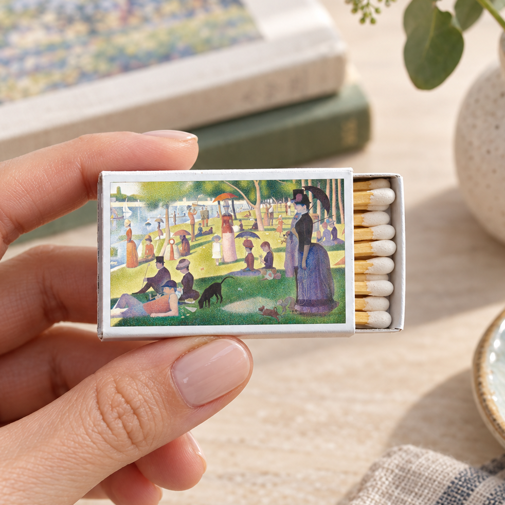 A hand holds a matchbox featuring a colorful impressionist painting of a park scene. The matchbox is open, showing white matches with gold tips, set against a softly blurred tabletop background with books and a vase.