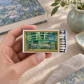 A hand holds a matchbox featuring a painted garden scene with a green bridge over water. The matchbox is shown in a styled tabletop setting with books, a vase, and fabric in the background.