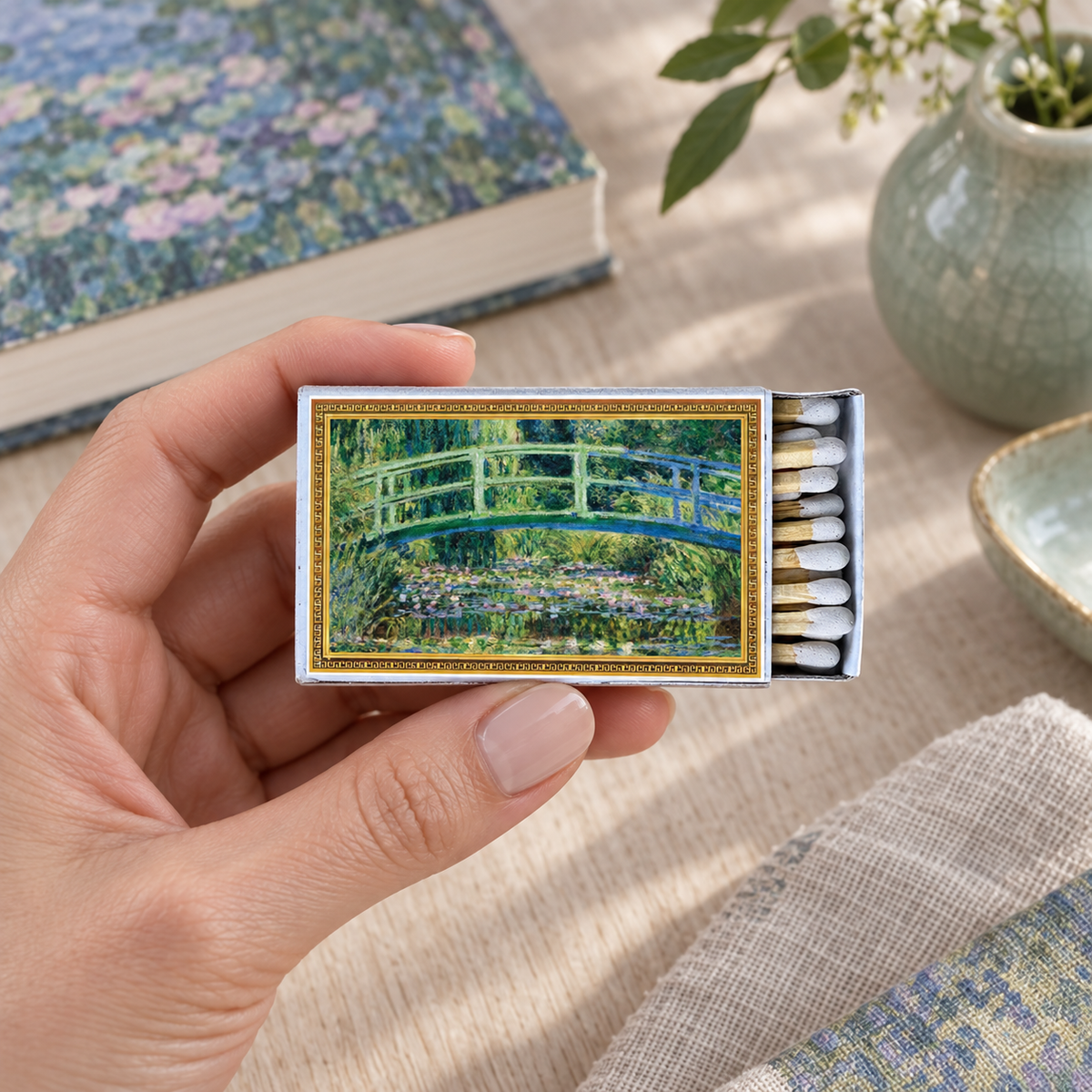 A hand holds a matchbox featuring a painted garden scene with a green bridge over water. The matchbox is shown in a styled tabletop setting with books, a vase, and fabric in the background.