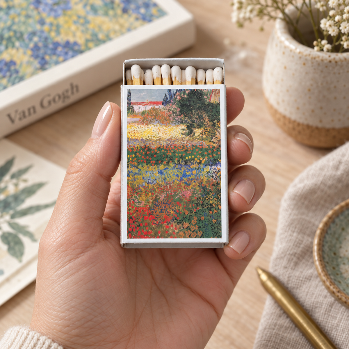 A hand holds a matchbox decorated with a colorful impressionist flower painting. The matchbox is open to show white matches inside, set against a styled tabletop background with a book, ceramic vase, and linen cloth.