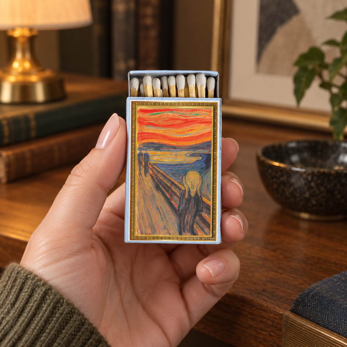 A hand holds a matchbox featuring Edvard Munch's painting 'The Scream' in a cozy, warmly lit room with books and a lamp in the background.