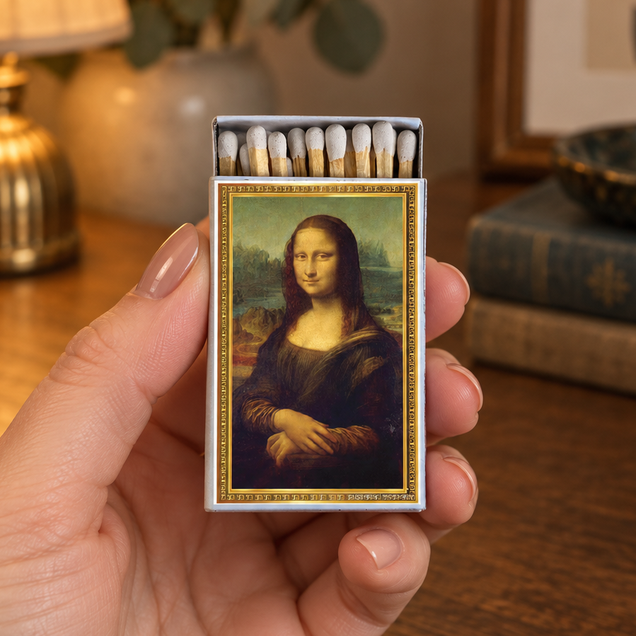 A hand holds a matchbox featuring a reproduction of Leonardo da Vinci's Mona Lisa painting, with match heads visible in the open top. The background shows a warm, cozy, blurred desk scene with a lamp, books, and a plant.