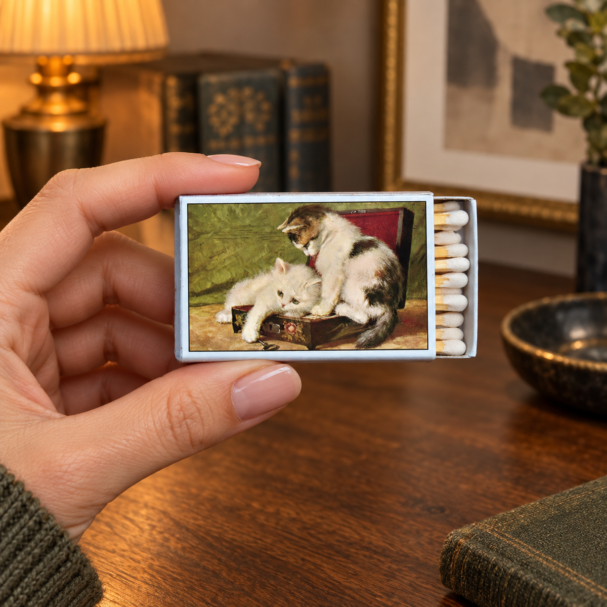 A hand holds a matchbox with a vintage-style print of two kittens in a box, set against a warm, cozy background with a lamp, books, and a wooden table.