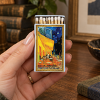 A hand holds a matchbox featuring Vincent van Gogh's 'Café des Artistes' painting. The matchbox is shown in a cozy, warmly lit indoor setting with books and a lamp in the background.