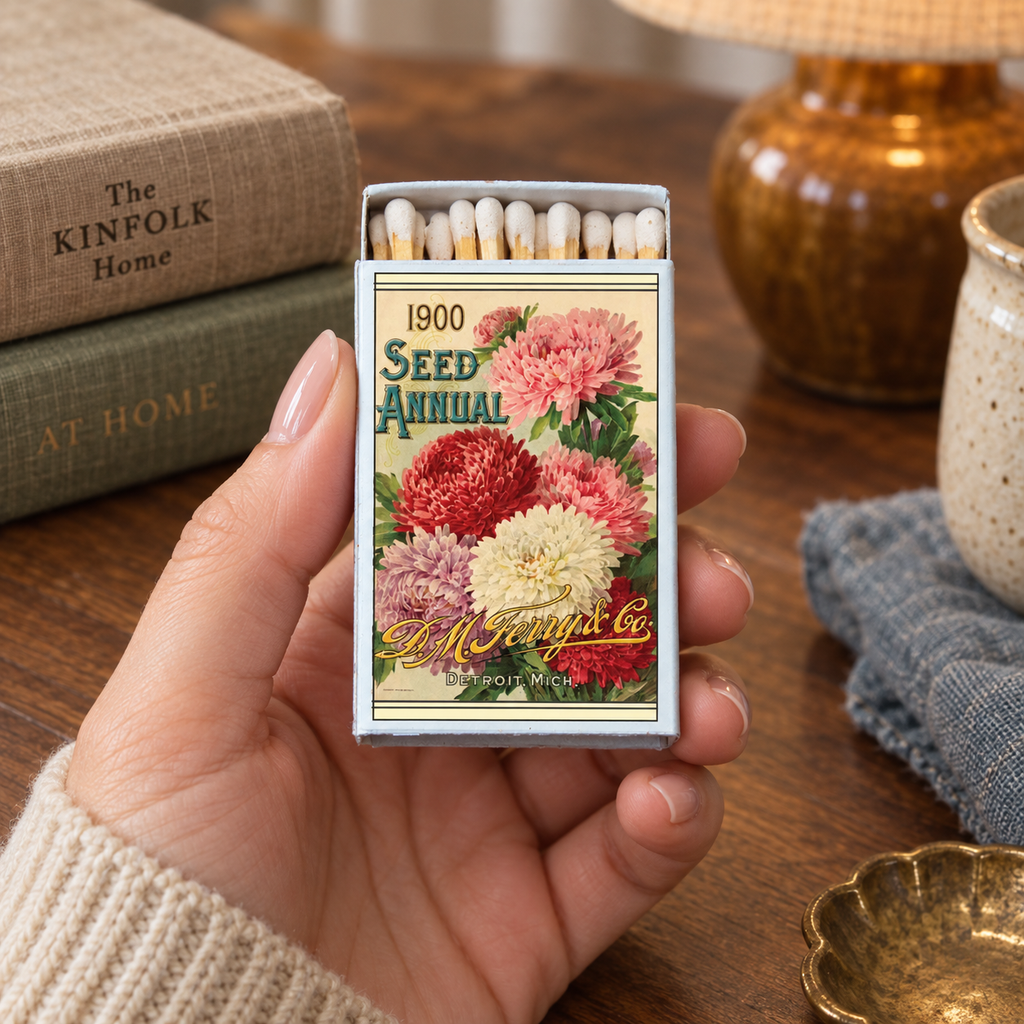 A hand holds a decorative matchbox shaped like a vintage 1900 seed annual book cover, showing colorful floral artwork and exposed matches inside. The scene is set on a wooden table with books, a lamp, and a small dish in the background.