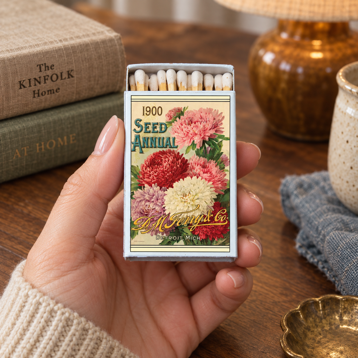 A hand holds a decorative matchbox shaped like a vintage 1900 seed annual book cover, showing colorful floral artwork and exposed matches inside. The scene is set on a wooden table with books, a lamp, and a small dish in the background.