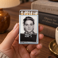 A hand holds a miniature matchbox-style lighter featuring a black-and-white portrait of Elvis Presley with the text 'PRESLEY ELVIS' on the front. The lighter is shown in a warm, styled home setting with a lamp, books, and a cup in the background.