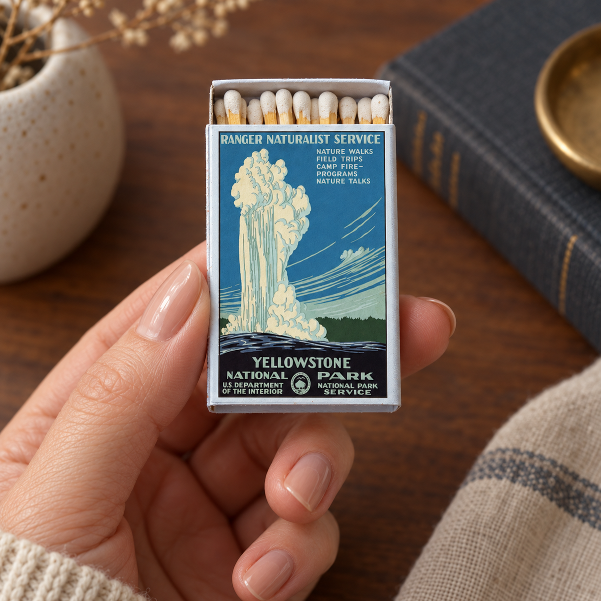 A hand holds a vintage-style Yellowstone National Park matchbox with matches visible inside. The box features a blue and cream illustration of a geyser eruption, with text reading 'Yellowstone National Park' and 'Ranger Naturalist Service.