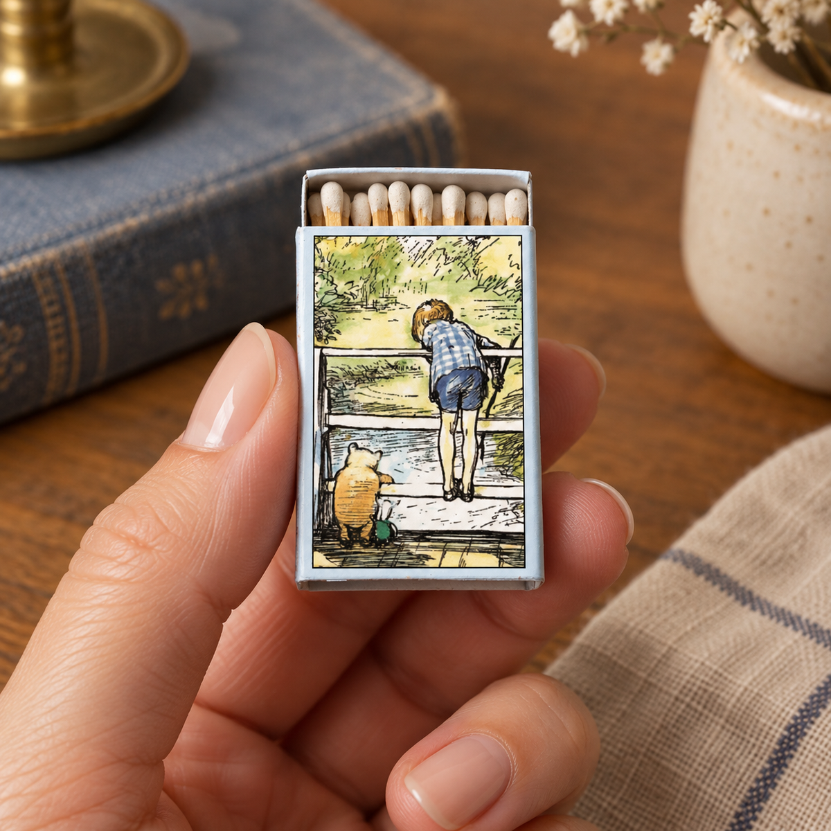 A hand holds a miniature matchbox with a detailed illustration of a child standing by a river, set against a warm, rustic background with a blue book and a ceramic vase.