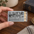 A hand holds a matchbox with a black-and-white illustration of three dancing skeletons. The matchbox is open, showing white matches with yellow tips, set on a wooden table with a vintage book and other decor in the background.