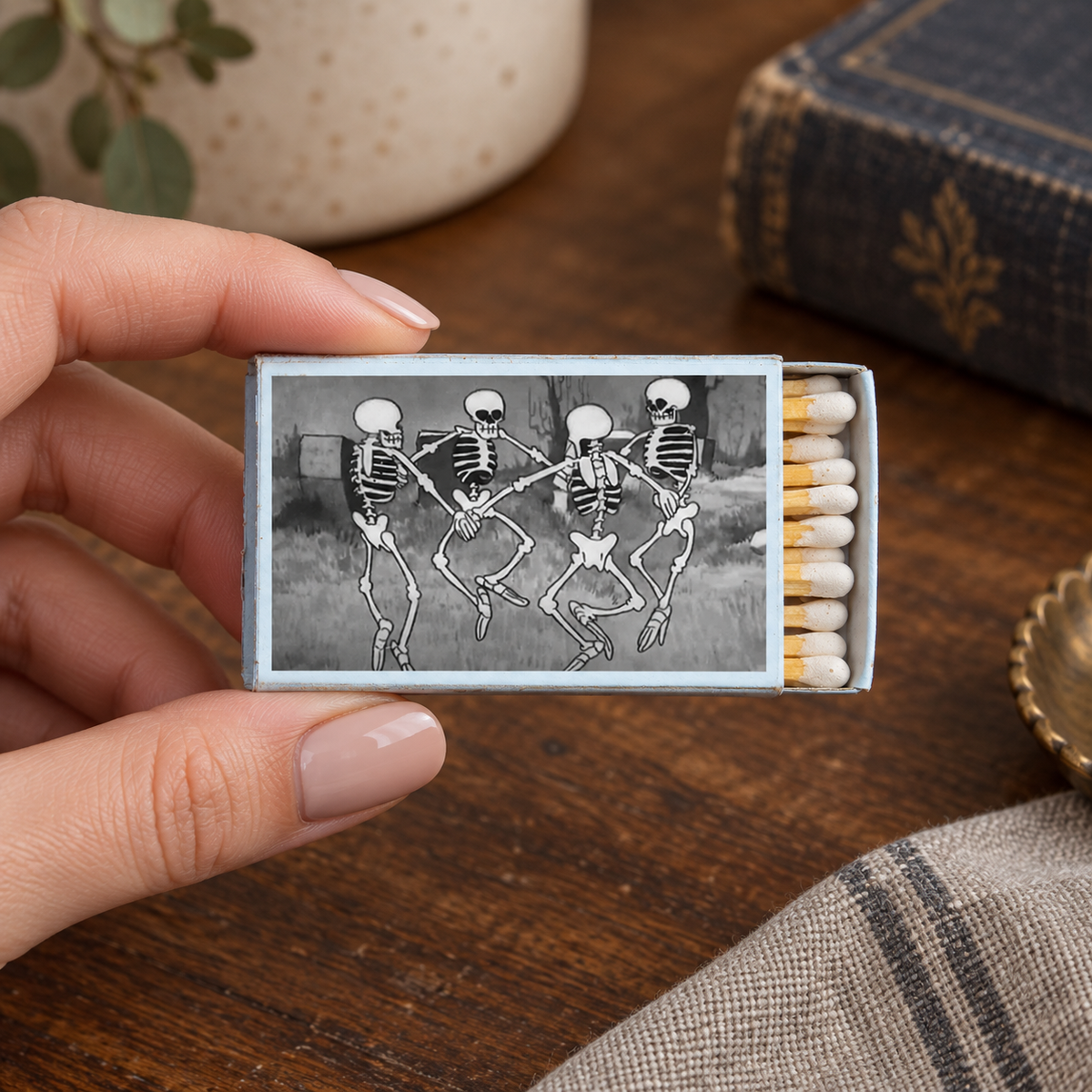A hand holds a matchbox with a black-and-white illustration of three dancing skeletons. The matchbox is open, showing white matches with yellow tips, set on a wooden table with a vintage book and other decor in the background.
