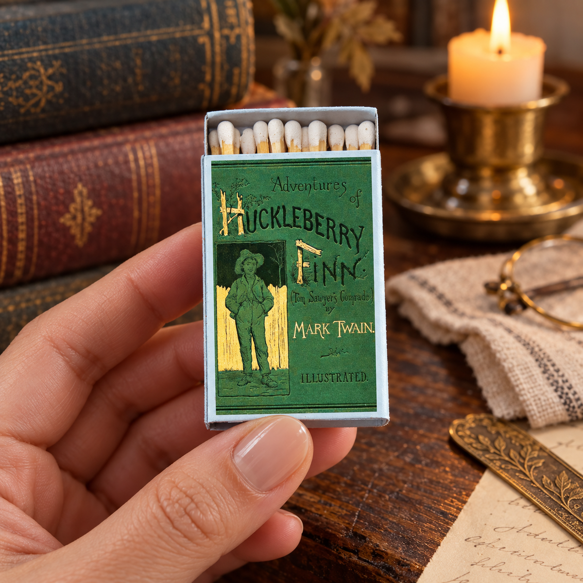 A hand holds a vintage-style matchbox featuring the cover art for 'Adventures of Huckleberry Finn' by Mark Twain. The matchbox is open, showing white matches inside, and is set against a warm, rustic background with a lit candle, old books, and paper.