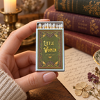 A hand holds an open vintage-style matchbox labeled 'Little Women' with white matches inside. The background shows a warm, styled scene with old books, a candle, and a piece of paper with floral details.