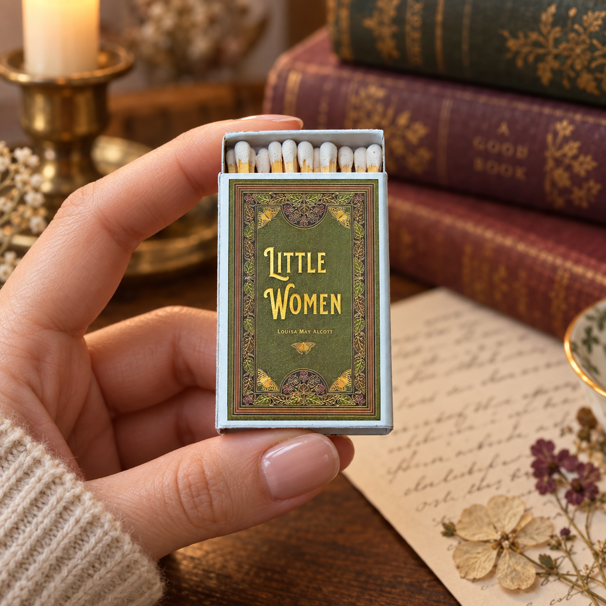 A hand holds an open vintage-style matchbox labeled 'Little Women' with white matches inside. The background shows a warm, styled scene with old books, a candle, and a piece of paper with floral details.