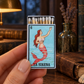 A hand holds a miniature matchbox-style lighter shaped like a La Sirena tarot card, with wood-toned matches visible in the open top. The background is a styled vintage desk scene with books, a lamp, and a globe.