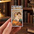 A hand holds a decorative matchbox featuring an Art Nouveau illustration of a woman in an orange dress against a gold and cream circular background. The scene is set on a rustic wooden desk with a brass lamp, books, and stationery in the background.