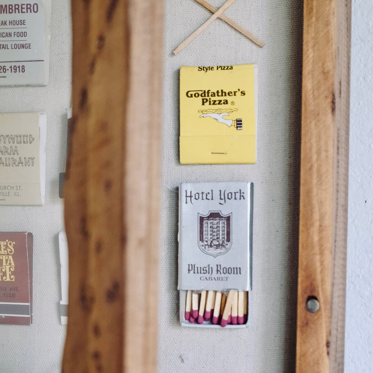 A close-up of a vintage wooden display case or cabinet with a light gray interior. Inside, small matchboxes are mounted as decor, including a yellow 'Grandfather's Pizza' matchbox and a white 'Hotel York' matchbox, with two wooden matches crossed above.