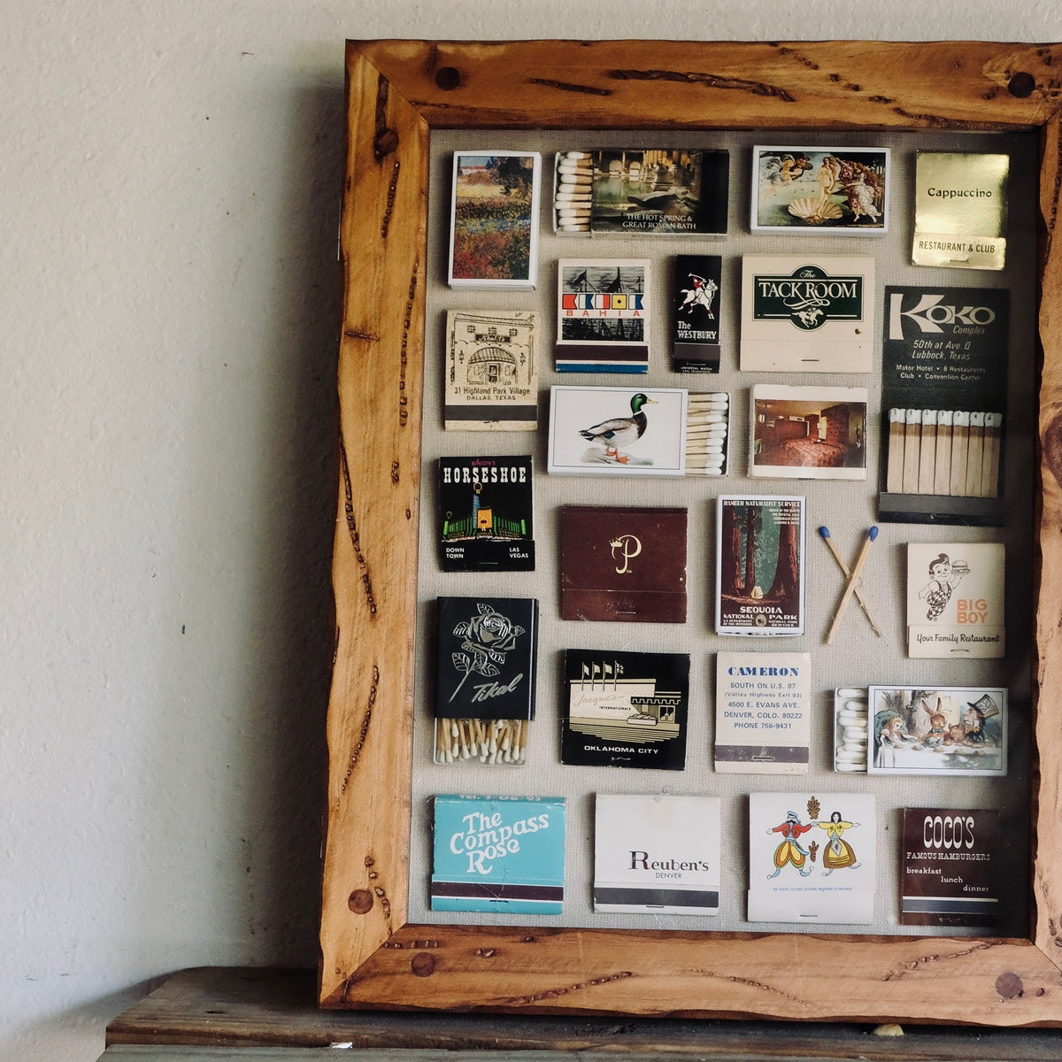 A rustic wooden shadow box frame filled with small vintage matchbooks, cards, and decorative pieces, displayed leaning against a white wall on a weathered wooden surface.