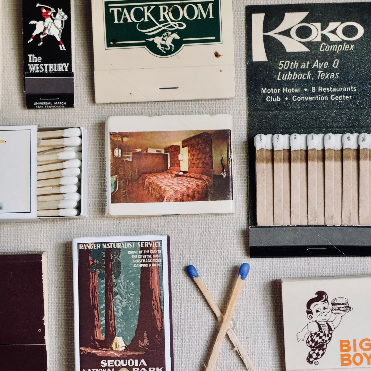 A collage of vintage matchbook covers, matchbooks, wooden matches, and small printed cards arranged on a light fabric background. Visible text includes 'The Tack Room,' 'Koko Complex,' 'The Westbury,' 'Big Boy,' and 'Cameron.