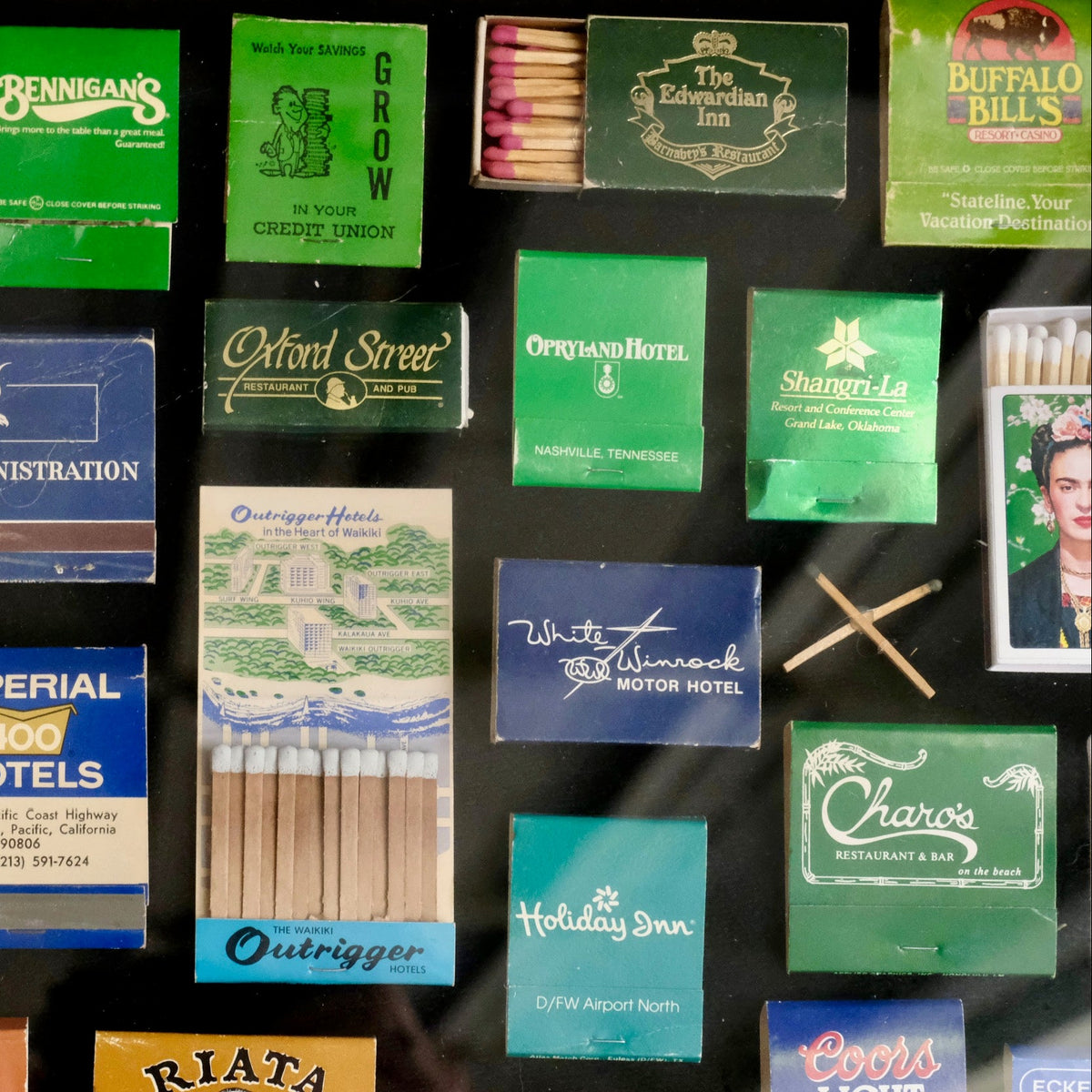 A collage of vintage matchboxes and cigarette packs in assorted colors, including green, blue, pink, and orange, arranged on a dark background. Visible brands include Imperial 400 Motels, Outtrigger, Chex, and Buffalo Bills.