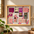 A framed collage of vintage matchboxes, matchsticks, and printed cards arranged inside a light wood shadowbox frame, hung on a beige wall above a wooden surface with plants and decor visible.